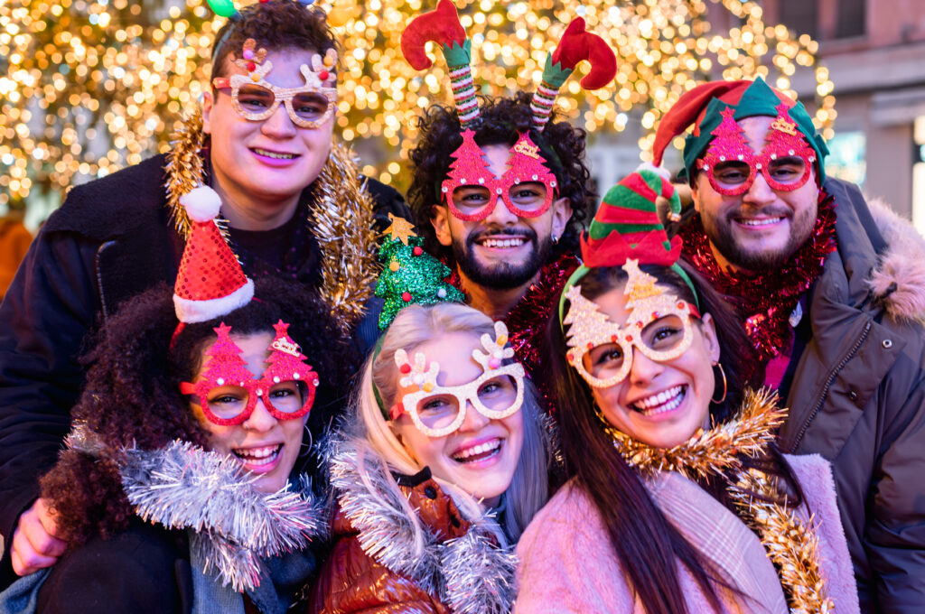 Group of friends wearing festive holiday glasses and accessories, smiling and celebrating together at a themed holiday party.